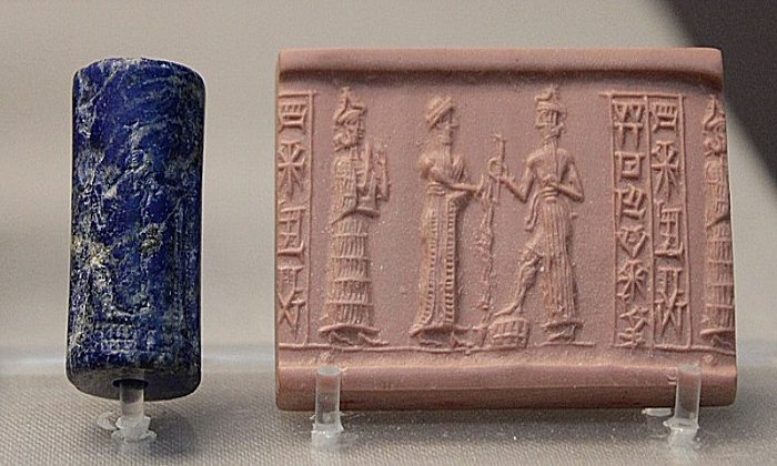 Lapis lazuli cylinder seal. A suppliant goddess (left, with necklace counterweight) stands behind the robed king (center) who pours libation before the ascending sun god who holds the rod and ring of justice and rests his foot on a rectangular chequer-board mountain. Bur-Dagan, son of Kurub-Adad, inscribed on the seal, is probably the name of the owner. From a tomb in Ur, dated c. 1900 BC. BM 121418.