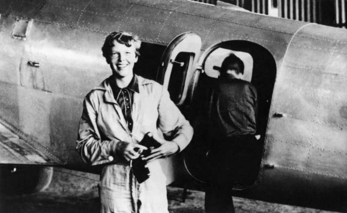 American aviator/pilot Amelia Earhart (1897-1937) standing by her Lockheed Electra dressed in overalls, with Fred Noonan getting into the plane in the background.